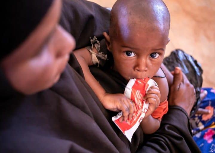 Le 3 février 2022 en Somalie, un enfant se nourrit d'un paquet d'aliments thérapeutiques prêts à l'emploi tandis que sa mère le tient dans ses bras en attendant de recevoir de l'aide au centre de santé Community Empowerment and Development Action à Dolow. ©UNICEF/UN0591078/Taxta
